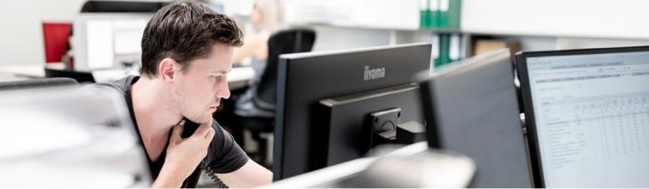 In einer Seitenansicht ist ein Mann zu sehen, der vor einem Computer sitzt. Der Mann mit kurzen dunklen Haaren trägt ein schwarzes Oberteil. Seine Hand ist in der Nähe seines Gesichts erhoben und erweckt den Eindruck von Nachdenklichkeit. Vor ihm zei