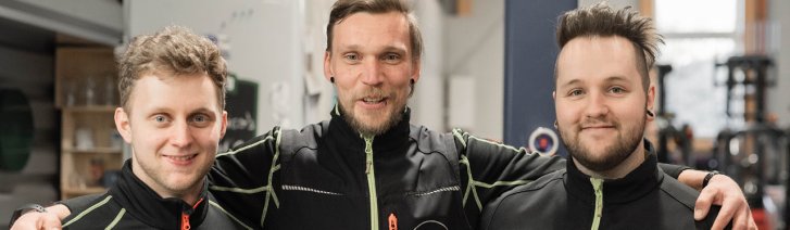 Drei kaukasische Männer in schwarzen Jacken, wahrscheinlich Teamkollegen oder Kollegen, stehen eng beieinander und haben die Arme verschränkt. Der Mann in der Mitte ist etwas größer als die anderen und scheint Bartstoppeln zu haben. 