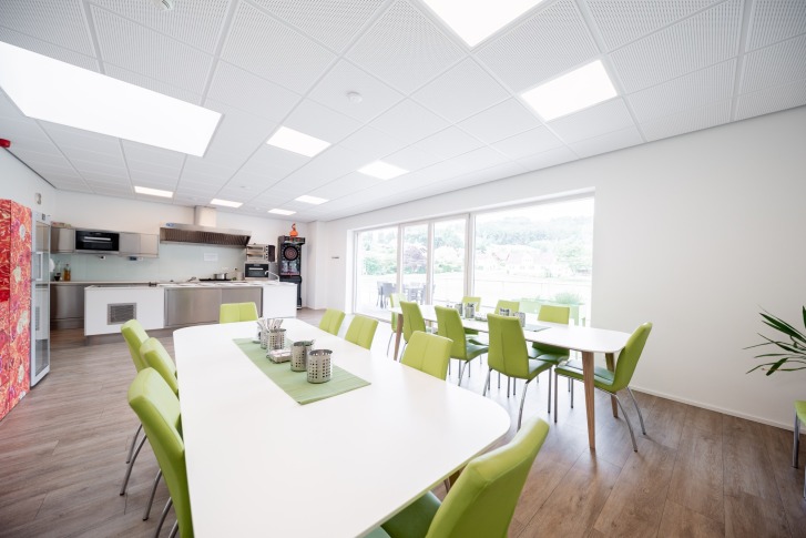 Lichte kamer met groene stoelen, grote tafels, kitchenette en uitzicht naar buiten door de ramen.