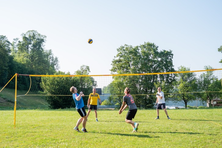 Four men play volleyball on a grass court with a net, surrounded by trees.