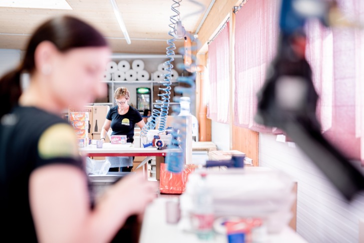 Two women pack products in a bright room with boxes and packaging material.