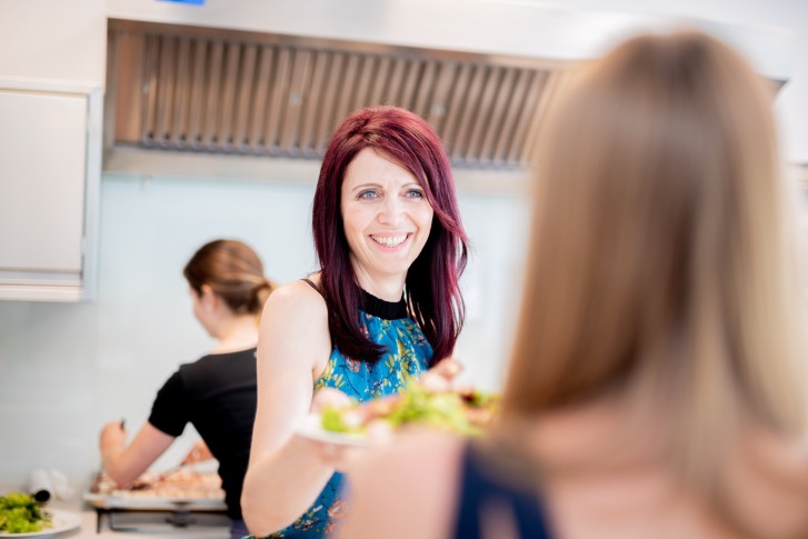 Woman smiling and passing plates to another person in a modern, bright kitchen.