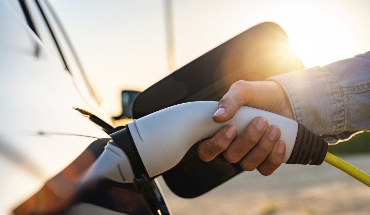 Hand met oplaadkabel aangesloten op auto buiten bij zonsondergang.