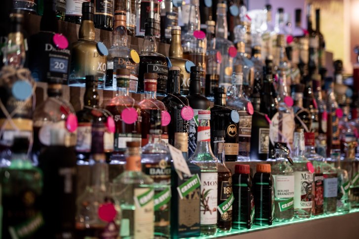 Een groot aantal flessen sterke drank staat op verlichte planken met kleurrijke prijskaartjes.