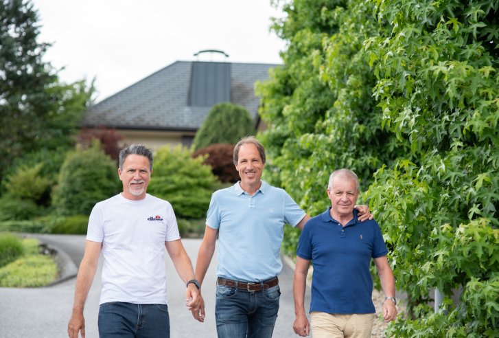 Drie mannen lachen in een magazijn met rekken vol dozen op de achtergrond.