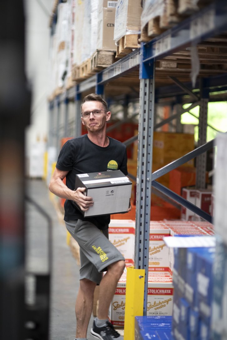 Ein Mann trägt eine Kiste durch ein Lager mit gestapelten Kartons auf Regalen.