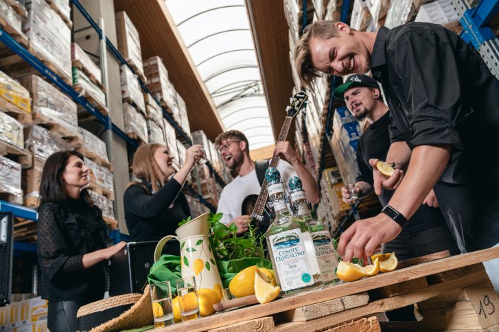 Mehrere Menschen stehen zusammen, lachen – im Hintergrund sind Flaschen mit Alkohol zu sehen.