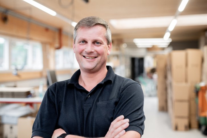 Ein Mann lächelt, steht mit verschränkten Armen in einem Lagerraum mit Kartons.