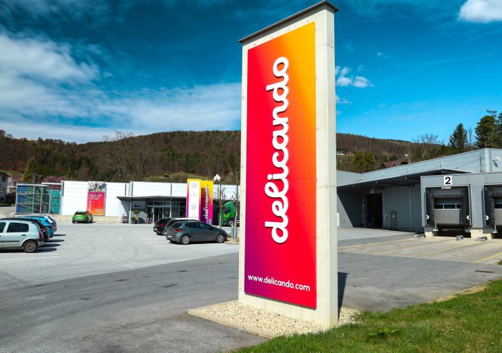 Une grande enseigne colorée avec le logo "delicando" se trouve devant un bâtiment moderne dans un environnement rural.