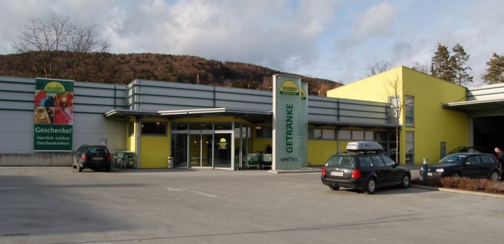 Gebouw staat rustig op parkeerplaats, omringd door auto's en bomen, met bord "GETRÄNKE".