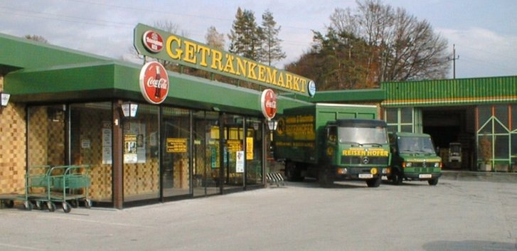 Getränkemarkt, grüne LKWs parken vor dem Gebäude. Werbung für Coca-Cola. Bäume im Hintergrund.