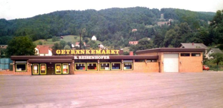 Beverage store building displays signs, stands in parking lot surrounded by wooded hills and residential homes.