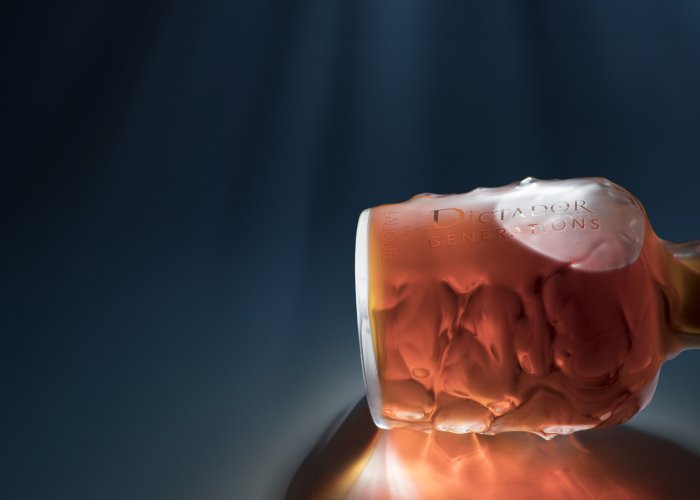 Eine edle Flasche mit rötlich-brauner Flüssigkeit liegt beleuchtet auf reflektierendem Untergrund.