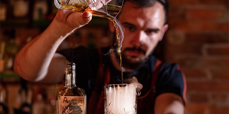 Man pouring drink into glass, bar environment, bottle with label "Animo" next to it.