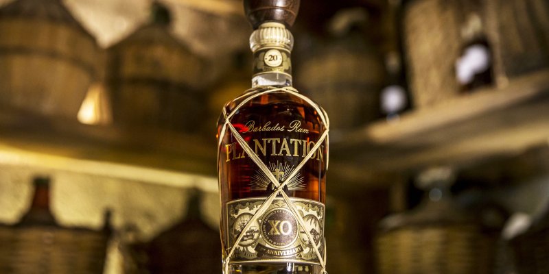 La bouteille de rhum est posée sur un tonneau en bois, entourée de paniers, dans un entrepôt rustique.