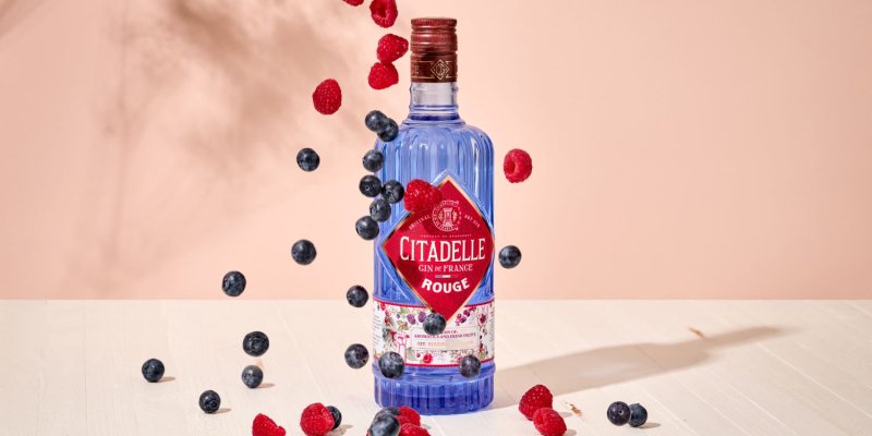 Eine blaue Gin-Flasche umgeben von schwebenden Heidelbeeren und Himbeeren auf hellem Holztisch.