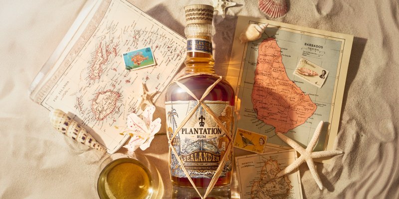 Rumflasche auf Sand mit Karten, Muscheln und einem gefüllten Glas daneben.