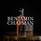 Flasche Benjamin Chapman Whiskey steht auf goldenem Tisch, schwarzer Hintergrund.