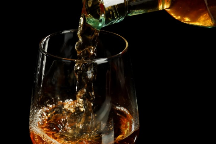 Whiskey wird in ein Glas mit Eiswürfeln gegossen, weitere Gläser im Hintergrund.