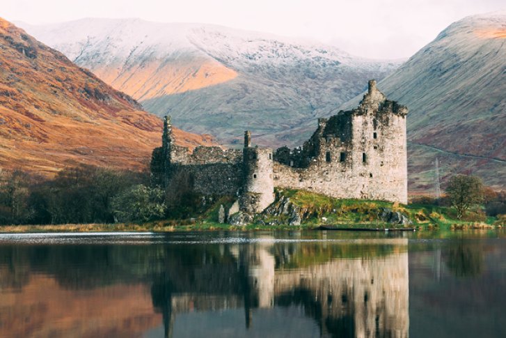 Eine Burgruine spiegelt sich ruhig im See, umgeben von Bergen und herbstlicher Landschaft.