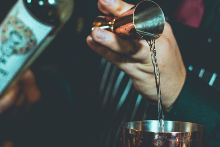 Hände gießen alkoholisches Getränk aus Flasche durch Messbecher in Metallbecher, in Barumgebung.