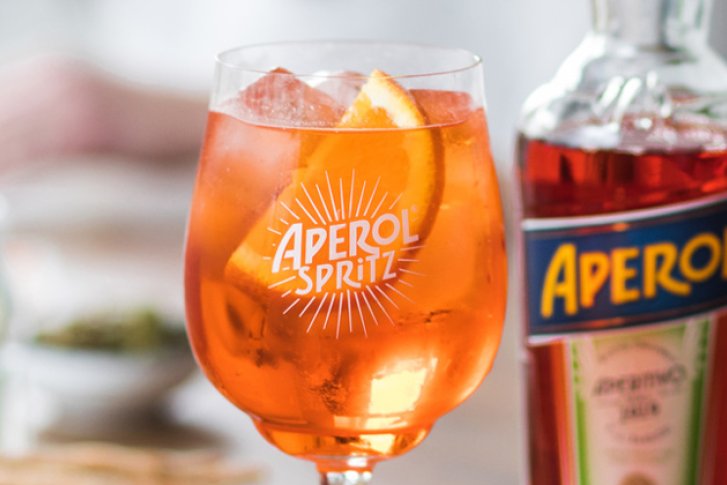 Ein Aperol-Spritz-Glas steht gefüllt neben einer Aperol-Flasche auf einem gedeckten Tisch.