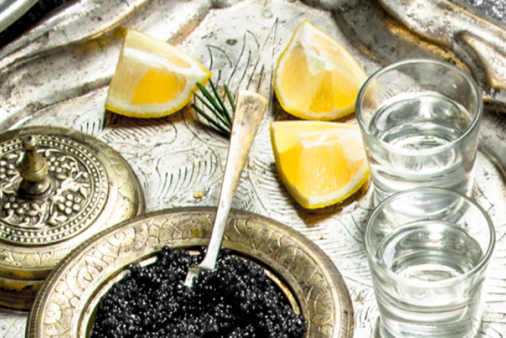Silbertablett mit Wodkagläsern, Kaviar, Zitronenscheiben und Dekor, auf rustikalem Untergrund.