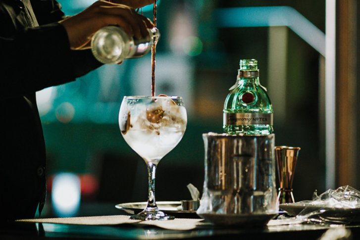 Barkeeper füllt Cocktailglas mit Eiswürfeln, Barhintergrund mit Flasche und Utensilien.