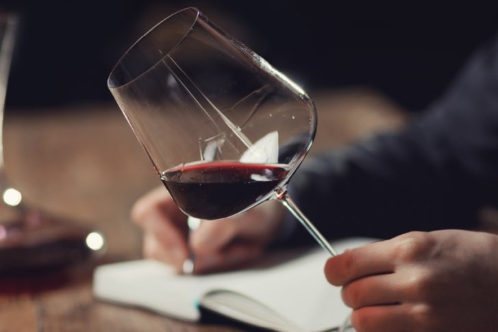 Weinglas kippen Hand, Dekanter und Notizbuch auf Holztisch, dunkler Hintergrund.