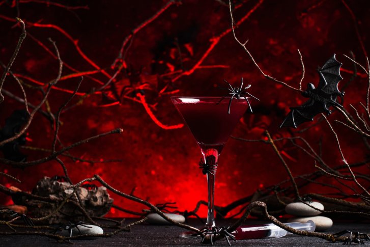 Ein rotes Cocktailglas steht zwischen Zweigen, dekoriert mit Kunststoff-Spinnen und einer Fledermaus.
