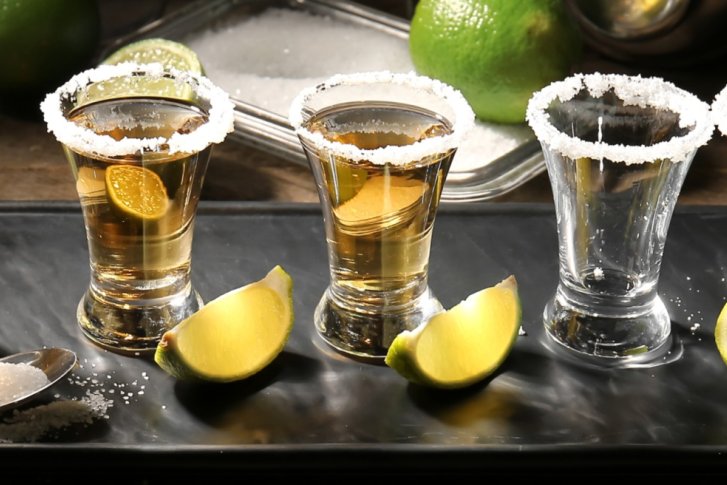 Fünf Tequila-Gläser mit Salzrand stehen auf einem Tablett, umgeben von Limetten.