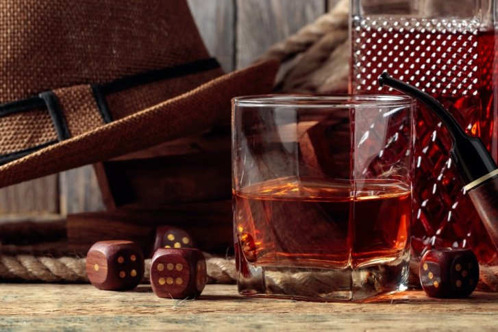 Glas Whisky neben einer Pfeife und Hut auf Holztisch vor rustikalem Hintergrund.