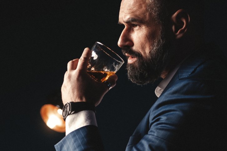 Mann hält ein Glas Whiskey, sitzt nachdenklich in schwach beleuchtetem Raum.