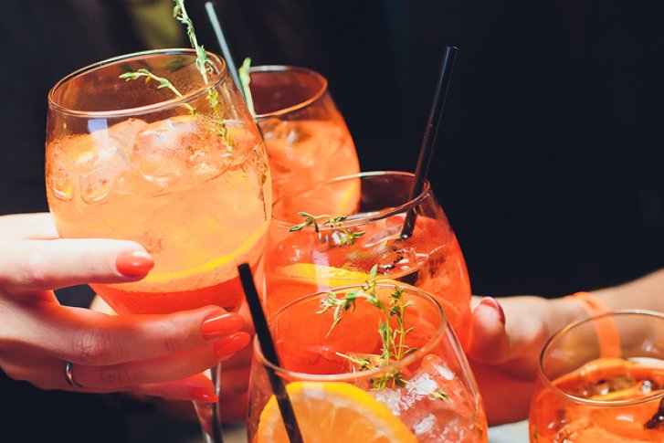 Gläser mit Aperol-Spritz stoßen an, umgeben von Händen und dunkler Hintergrundbeleuchtung.