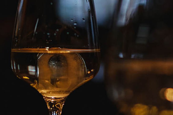 Weinglas mit goldenem Getränk steht vor dunklem, unscharfem Hintergrund in stimmungsvoller Beleuchtung.