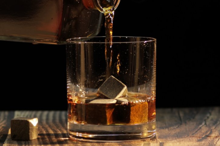 Glas mit Eiswürfeln, Getränk wird eingeschenkt, auf einem Holztisch vor schwarzem Hintergrund.