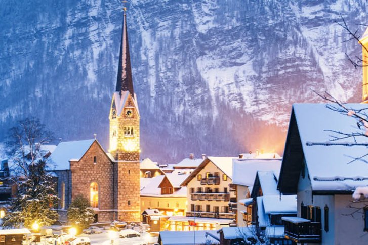 Verschneiter Kirchturm ragt aus einer malerischen, beleuchteten Dorflandschaft vor verschneiten Bergen empor.