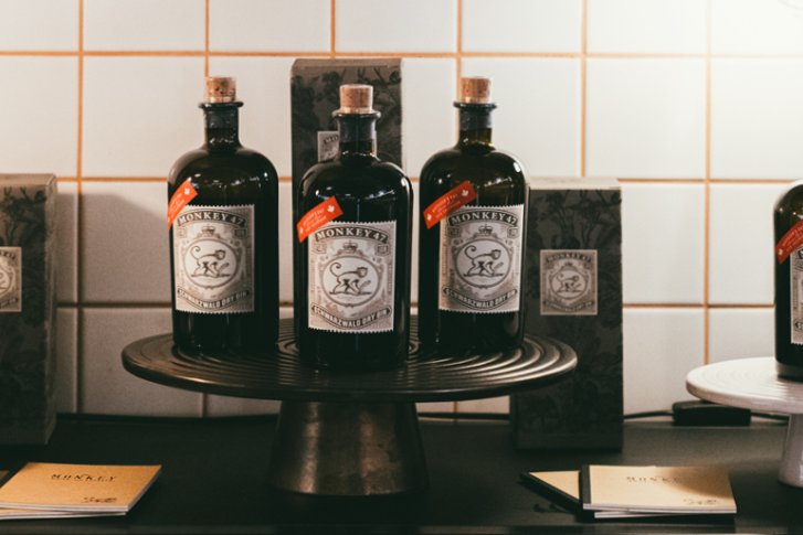 Flaschen stehen auf Podesten vor Kacheln, umgeben von Schachteln, mit "Monkey 47" Etiketten.