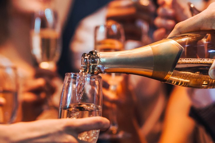 Flasche Sekt wird in Gläser gegossen, umgeben von Menschen in festlicher Atmosphäre.