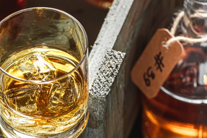 Ein Glas mit goldener Flüssigkeit, steht mit Eiswürfeln, neben dekorativen Flaschen im Holzregal.