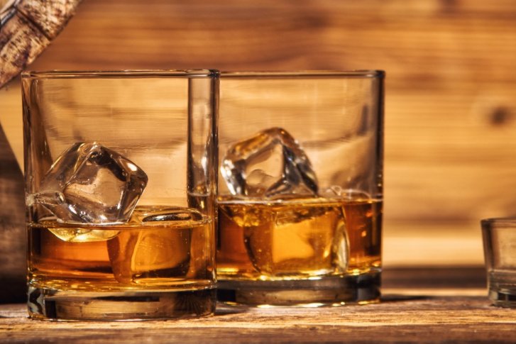 Zwei Whiskeygläser mit Eis stehen neben brennender Zigarre auf einem Holztisch mit Fass.