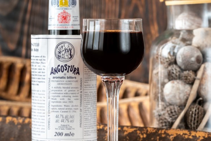 Glas dunkles Getränk steht vor Angostura-Flasche, daneben Dekoglas mit Steinen auf Holztisch.