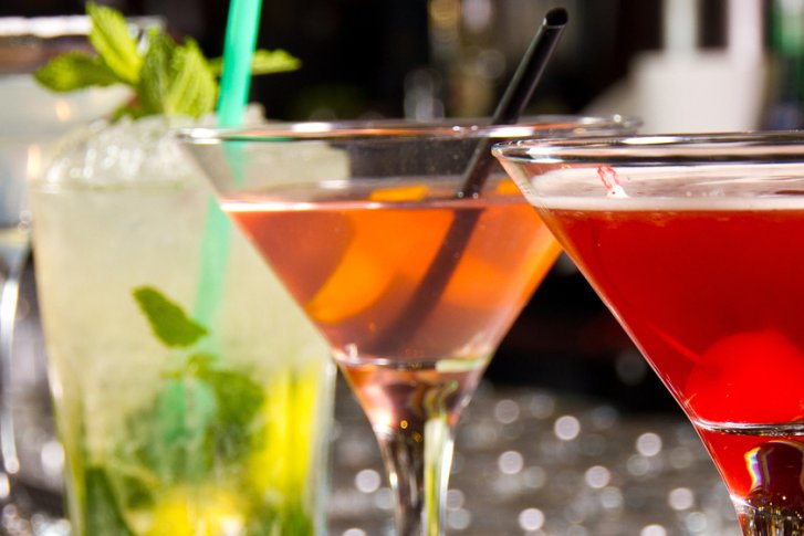 Verschiedene bunte Cocktails stehen nebeneinander auf einer Bar, teils mit Strohhalmen garniert.