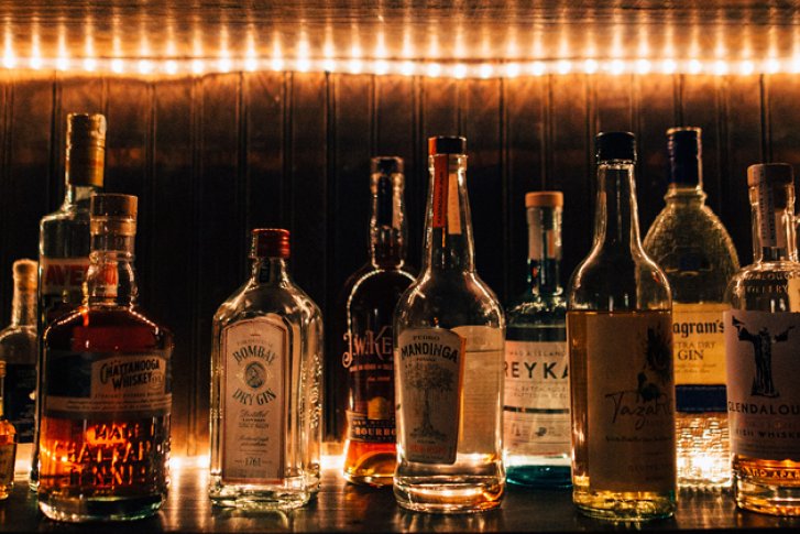 Verschiedene Spirituosenflaschen stehen beleuchtet auf einem Regal aus Holz in dunkler Umgebung.