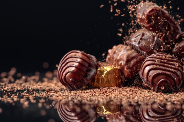 Pralinen liegen gestapelt, umgeben von Schokoladenstreuseln, auf einer reflektierenden, dunklen Oberfläche.