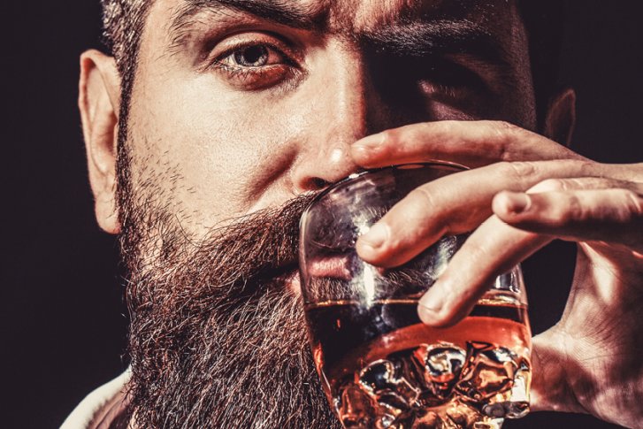 Mann hält ein Glas mit dunklem Getränk nahe am Gesicht, vor dunklem Hintergrund.