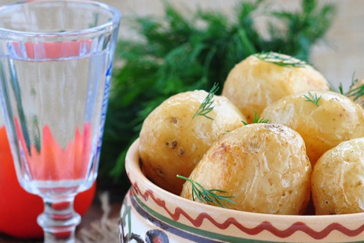 Kartoffeln im traditionellen Keramikteller mit Dill, begleitet von Schnapsglas, Tomate und Zwiebel auf Holztisch.