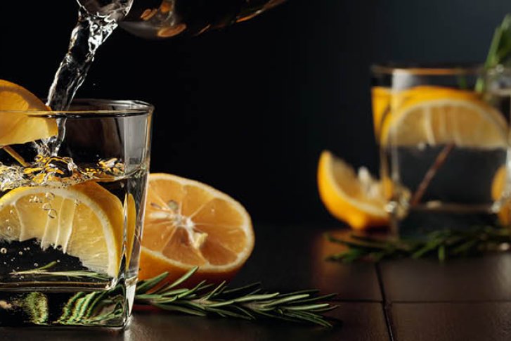 Glas wird mit Getränk gefüllt, enthält Zitronenscheiben und Rosmarin, auf dunklem Holztisch.