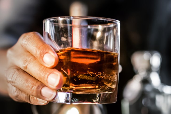 Eine Hand hält ein Whiskyglas, im Hintergrund sind Barutensilien unscharf zu erkennen.