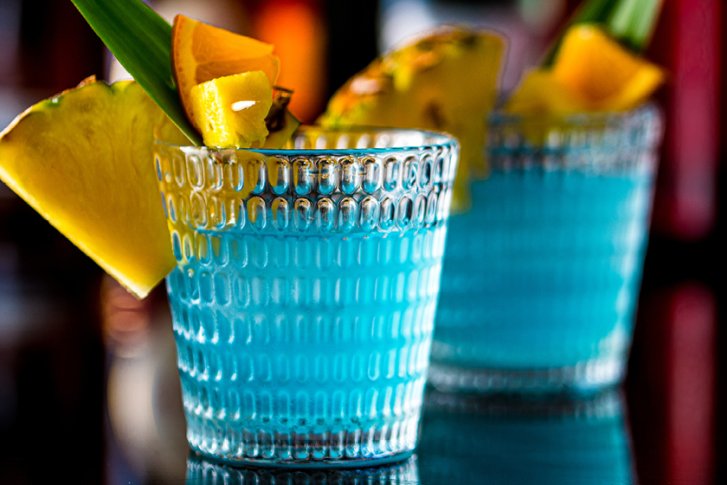 Blaue Cocktails mit Ananas und Blattgarnitur stehen auf schwarzem, reflektierendem Tischuntergrund.